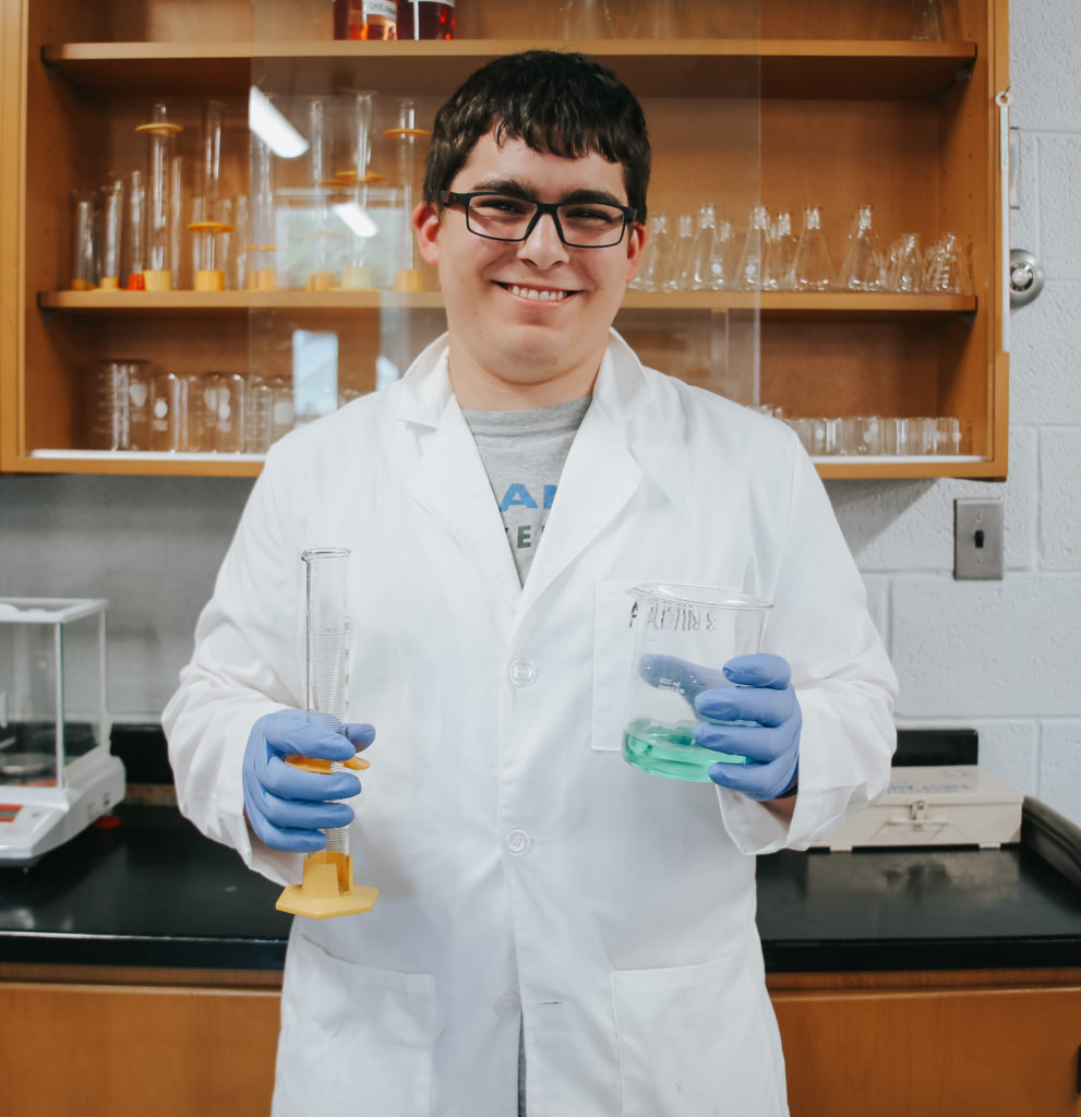 Austin Keranen wearing a white lab coat jacket, smiling, and holding two glass beakers used for measuring out liquids in the biology lab.
